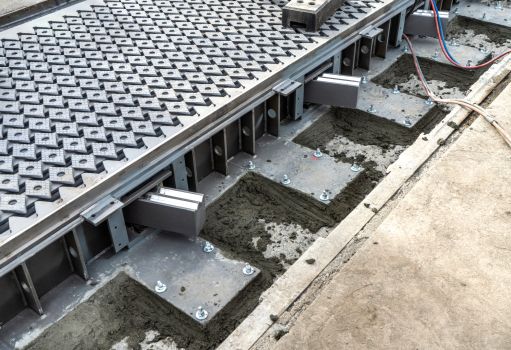 The expansion joint after installation : This image shows the joists with the gleaming W-shaped catamaran supports. These support the profiles running transverse to the road direction, with the rhomboid plates for noise re-duction on top.