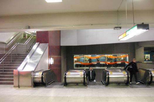 Dans les escaliers mécaniques, passagers accédants du niveau ligne Verte au niveau ligne Orange; l'escalier de granit à gauche conduit au niveau mezzanine. En arrière plan de l'autre côté des baies vitrées, un train est en station sur la ligne Orange. 13/27 ligne Orange métro de Montréal