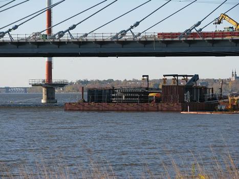 Les derniers éléments du pont arrivent sur barge. Remarquez ce que l'on verra en roulant vers Montréal; à gauche sur la ligne d'horizon, le pont Pie-IX et plus loin encore la centrale hydroélectrique Rivière des Prairies