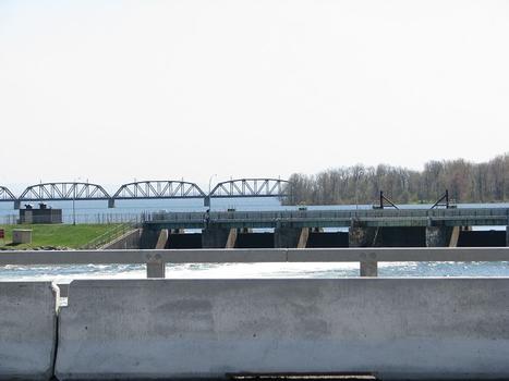 Grossissement au téléobjectif du pont ferroviaire des Coteaux qui est situé en arrière plan sur la ligne d'horizon. Au premier plan en bas, le parapet du pont Mgr Langlois, et entre les deux ponts, la digue d'Hydro Québec. En amont du pont ferroviaire, c'est le lac Saint-François, un élargissement du fleuve Saint-Laurent qui s'étend jusqu'à Cornwall en Ontario
