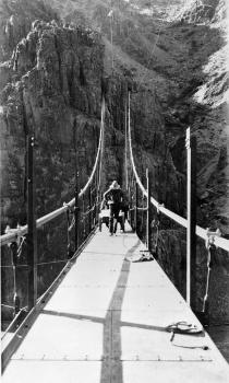 Kaibab Trail Suspension Bridge: Transporting steel floor plates on hand cart to north end of bridge. Note retaining angle for surfacing at cut-side edge of floor plates. Note tunnels in top of rock face for main cables 
(HAER, ARIZ,3-GRACAN,3-22)