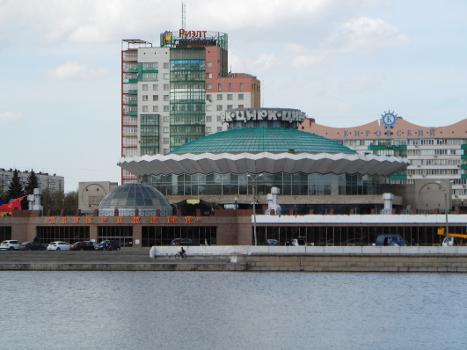 Circus building at Kirova Street, 25 in Chelyabinsk : View from the opposite right bank of the Miass River (ChGRES pond). South facade of the building. In the foreground is the shopping center Naberezhny (Kirova street, 27), in the background the building of the residential complex Kirovsky (Kirova Street, 23a and Brat'yev Kashirinykh Street, 12).