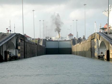 One of the lock chambers in the Panama Canal. The view is from a small boat about to enter the east flight of the Gatun locks, heading south towards the Pacific side. A ship can be seen in the next chamber up; on either side of the lock are the railway tracks on which the mules run. Within the arches to either side in the foreground are the halves of a swinging road bridge