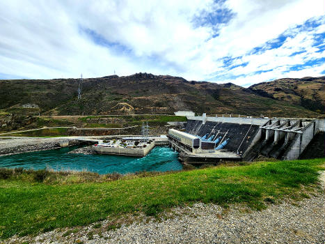 A picture of the Clyde Dam in September 2022, Otago, New Zealand, taken from a slight medium-distance depicting the outlet side of the dam, surrounding terrain, and the platform where the administration buildings for the dam are located.