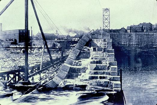 Holyoke Dam : A cross section photograph of the Holyoke Dam during the third and present structures construction in the late 1890s, showing the combined inner masonry and outer granite cap. The tower in the background is one of two belonging to once the largest cableway in the world, used for transporting some materials along the height of the structure, in tandem with the narrow gauge rail (seen left) and several cranes.