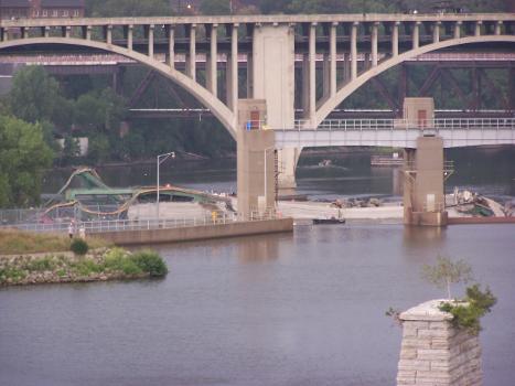 Collapsed I-35W bridge in Mississippi River, August 1, 2007. Two rescue boats are visible through dam arch, with concrete carts near cars on bridge deck. View is from Stone Arch Bridge across lower St. Anthony Falls dam, with 10th Street bridge in background