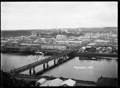 Panoramic view of Wanganui across the Whanganui River:Cobham Bridge is in the foreground, with the business premises of M. Hogan &amp; Company Ltd., H.I. Jones &amp; Sons Ltd., and the Wanganui branch of the New Zealand Loan &amp; Mercantile Agency Company, on the far side of the river. Photograph taken by William Archer Price. 
Inscriptions: Inscribed - Photographer's title on negative -bottom right: Wanganui.