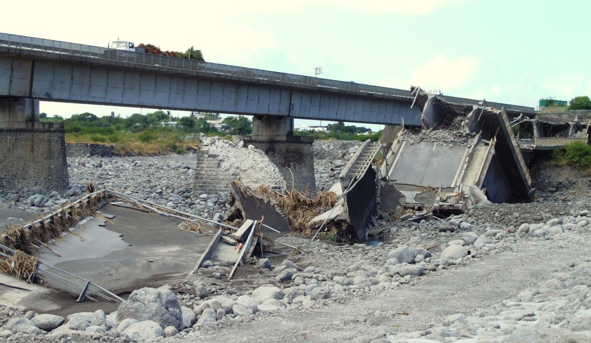 Fiche média no. 81618 Saint-Pierre - Le pont aval de la rivière Saint-Etienne effondré. Une pile a été cisaillée transversalement à cause du mouvement du tablier vers l'aval. Cette chute sur le côté a entrainé un mouvement longitudinal du tablier vers le centre du pont et conduit à une rupture de la pile suivante