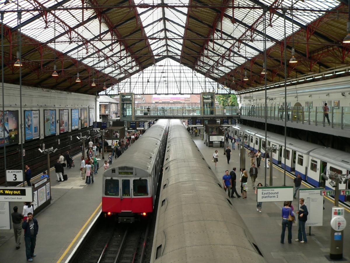 The District Line platforms at Earl's Court London Underground station viewed from the main concourse. The leftmost train is an unrefurbished London Underground D78 Stock on a westbound service. The train on the far right platform is one of the refurbished D78 stock units on an eastbound service, that the platform indicators suggest is en-route to Tower Hill. The train in the centre of the picture is a London Underground C69 Stock train heading east to Edgware Road via High Street Kensington.