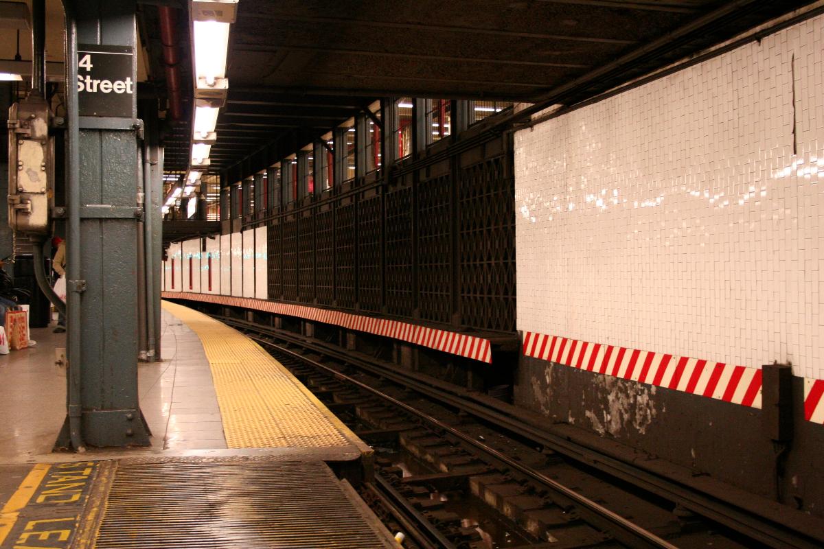 14th Street – Union Square Subway Station (Lexington Avenue Line) Local-Bahnsteig im U-Bahnhof an der IRT Lexington Avenue Line, Blickrichtung Süden. Hinter der rechten Wand und den Lüftungsgitter befindet sich ein ehemaliger Seitenbahnsteig, dessen Bahnsteigkante noch deutlich sichtbar ist. Darüber ist das Zwischengeschoss zu erkennen. Die weiß-roten Baken an der Wand weisen darauf hin, dass kein Raum zwischen Zug und Wand verbleibt. Im Vordergrund sind die beweglichen Bahnsteigkanten zu sehen, die über Lichtschranken im Gleisbett gesteuert werden. Der Schriftzug lautet „Stand ©le(ar)“, also „Zurückbleiben“.
