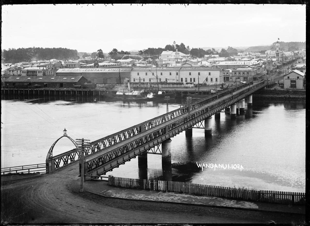 View of Wanganui across the Whanganui River Cobham Bridge is in the foreground, with the business premises of M. Hogan &amp; Company Ltd., H.I. Jones &amp; Sons Ltd., and the Wanganui branch of the New Zealand Loan &amp; Mercantile Agency Company, on the far side of the river. Photograph taken by William Archer Price.